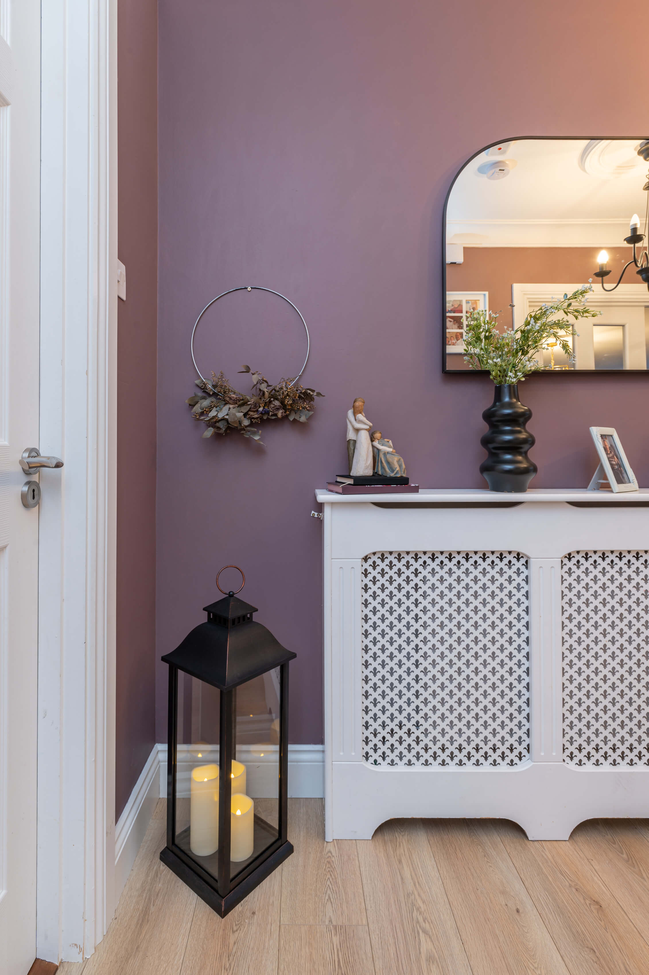 a corner of a hallway. Walls are a dusky purple colour, white radiator cover with ornaments, books and photos. There is a large black lantern with LED candles in the corner and a black arched mirror on the wall above the radiator. The floors are a light brown laminate.